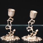Par de castiçais em prata, com receptáculos campanulares ornados por frisos verticais e folhagens em relevo, apoiados sobre hastes vazadas de volutas, com figura infantil em vulto pleno junto à base de formato oval recortada com decoração em relevo, estilo rococó. Medidas: Alt.: 10,5 x Comp.: 7 x Larg.: 5 cm. Peso: 240,7 gramas.