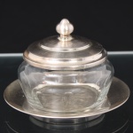 Elegante queijeira com recipiente em meio cristal, translúcido e incolor, formato circular, circundado por facetas de janelas ovais, fundo estrelado, tampa e presentoir em metal espessurado à prata. Medidas: Alt.: 9 x Diam.: 13 cm.