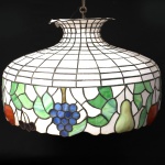 Luminária pendente em vitral, dito "tutti-frutti", composta por cúpula de formato bojudo com colo afunilado, estruturada por perfis metálicos e placas de vidro colorido. Decoração com motivos frutíferos, representando peras, maçãs, cerejas, cachos de uvas e folhagens estilizadas. Estilo Tiffany. Medidas: Alt.: x Diam.: Apresenta pequenas perdas e quebraduras na parte superior, vide foto.