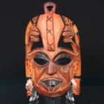 Máscara africana em madeira entalhada, formato antropomórfico com aberturas vazadas para olhos e boca, ornada por elementos escultóricos superiores representando figuras zoomórficas estilizadas. Decoração incisa e pintada em tons contrastantes, com reservas geométricas e motivos lineares. Provida de perfuração superior para suspensão. Medidas: Alt.: x Comp.: