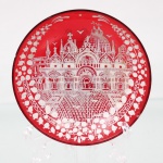 Prato de coleção em vidro veneziano vermelho translúcido, decorado em esmaltação branca com representação da Basílica de São Marcos, Veneza, circundada por friso de motivos vegetalistas estilizados. Corpo circular com pequeno pé anelar. Apresenta bicado na borda. Medida: Diam.: 10,5 cm.