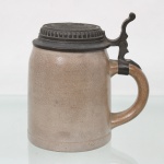 Tankard executado em grês vitrificado, de formato cilíndrico com ligeira curvatura superior, dotado de alça lateral e tampa articulada em metal fundido decorada em relevo com inscrição Franziskaner Bräu  Rio de Janeiro e representação de monge franciscano. Medidas: Alt.: 14,5 x Comp.: 13 x Larg.: 10 cm.