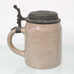 Tankard executado em grês vitrificado, de formato cilíndrico com ligeira curvatura superior, dotado de alça lateral e tampa articulada em metal fundido decorada em relevo com inscrição Franziskaner Bräu  Rio de Janeiro e representação de monge franciscano. Medidas:  Alt.: 14,5 x Comp.: 13 x Larg.: 10 cm.