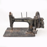 Máquina de costura manual Rainha, de acionamento por manivela lateral, confeccionada em ferro fundido esmaltado em preto, com decoração floral em policromia dourada aplicada por decalque. Base retangular metálica, volante raiado lateral e mecanismos aparentes. Inscrição Rainha na coluna frontal e De Máquinas de Costura na parte superior do braço. Exemplar de uso doméstico, produzido no início do século XX. Apresenta desgastes, pontos de oxidação e marcas de uso. Medidas: Alt.: 27 x Comp.: 49 x Larg.: 18,5 cm.