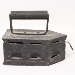 Ferro de passar à carvão em ferro fundido, marca Guarani, com tampa articulada e pega cilíndrica em madeira e metal, laterais vazadas em arcadas para ventilação do compartimento interno destinado ao carvão incandescente. Tampa móvel com sistema de fecho metálico lateral. Exemplar de uso doméstico, produzido no final do século XIX ou início do século XX. Medidas: Alt.: 17 x Comp.: 21 x Larg.: 10,5 cm.