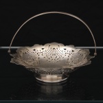 REGIA PRATA 90. Cesta de confeitos ou fruteira em metal prateado, de formato circular, interior côncavo, assente sobre base alteada e provida de alça móvel arqueada. Corpo executado em trabalho vazado e cinzelado, com decoração de volutas, elementos vegetalistas estilizados e fundos reticulados. Marca da manufatura no verso. Medidas: Alt.: 13 x Diam.: 17 cm. Apresenta marcas de uso e desgastes.