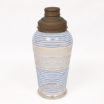 Coqueteleira (shaker) em vidro incolor, corpo cilíndrico levemente bojudo, decorado com faixas foscas alternadas por filetes dourados e azul. Tampa removível em metal com coador interno perfurado e sobretampa de encaixe. Medidas: Alt.: 23 x Diam.: 10 cm.