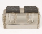 Tinteiro de mesa em bloco de cristal incolor, de formato retangular, com dois reservatórios internos para tinta e tampas quadradas em metal preto decoradas em relevo com motivos vegetalistas estilizados. Medidas: Alt.: 6,5 x Comp.: 13 x Larg.: 6,5 cm.
