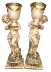 EUROPA - Pendant de magnificas floreiras em biscuit europeu representando figuras de querubins portando vasos com detalhes em vibrante ouro sobre base quadrangular com detalhes em guirlanda de flores. Mede 47cm.