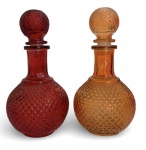 Par de garrafas decorativas em vidro moldado, uma em vermelho rubi e outra em tom âmbar, ambas com corpo bojudo e textura geométrica em relevo. Gargalos longos e tampas esféricas facetadas, conjunto elegante de forte apelo decorativo, com pátina do tempo. Mede 22 cm.