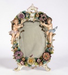 ALEMANHA SÉC. XIX - Magnifico espelho de parede com moldura em porcelana alemã do século XIX, ricamente ornada com flores em relevo policromadas e detalhes em douração. Composição decorada por dois putti em vulto, de modelagem delicada e pintura suave, sustentando guirlandas florais. Verso com suporte metálico original para fixação. Espelho com manchas do tempo e perdas consecutivas. Medidas: 33 x 24 cm. Peça de forte apelo decorativo, ideal para ambientações clássicas e colecionadores de bom gosto.