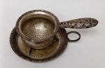 Coador de Chá Antigo com Descanso  Estilo Vitoriano  Prata / Metal TrabalhadoCoador de chá individual com base de gotejamento original.Cabo: Rico trabalho vazado com motivos de arabescos e volutas.Coador: Formato de taça com três pés de apoio para encaixe firme na base ou na xícara.Base: Pratinho com bordas cinzeladas e gravação floral no centro.