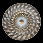 (LALIQUE) Importante centro de mesa, peça de autoria de René Lalique, executado em vidro moldado opalescente, apresentando exuberante decoração geométrica em relevo radial, de excepcional elegância e forte influência Art Déco.Peça assinada na parte inferiorda base, destacando a célebre técnica desenvolvida por Lalique, marcada pela interação entre transparência, opalescência e refinado trabalho escultórico da superfície.O modelo evidencia o virtuosismo criativo do mestre vidreiro francês, cuja obra revolucionou o design decorativo do século XX, tornando-se referência mundial em vidro artístico e alta decoração.