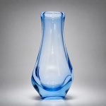 SANDRO ROSIN  Vaso em vidro artístico soprado à bocaElegante vaso em vidro artístico soprado à boca, atribuído ao designer Sandro Rosin, executado em delicada tonalidade azul transparente, de refinado acabamento e linhas orgânicas contemporâneas.A peça apresenta corpo alongado de perfil fluido, com base facetada e suaves movimentos internos do vidro, evidenciando o virtuosismo técnico característico da produção artesanal em vidro soprado. A transparência azulada, associada à pureza formal do desenho, confere leveza visual, sofisticação e forte presença escultórica.Exemplar representativo do design em vidro contemporâneo, conciliando expressão artística, funcionalidade e elevado apelo decorativo
