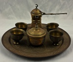 Conjunto oriental para em metal dourado, composto por bandeja circular ricamente gravada, pequena cafeteira de bico longo, quatro copinhos e açucareiro tampa, todos ornamentados com delicados motivos geométricos e arabescos de inspiração islâmica/mourisca. Medindo respectivamente: 31 cm, 16 cm, 10 x 9 cm e 5,5 x 6 cm.