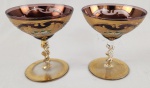 Par de taças em vidro veneziano na tonalidade lilás com rica decoração em ouro e detalhes de flores policroma. Haste espiral e pés com vestígio de ouro. Medindo: 13 x 12 cm.