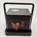 Elegante caixa em laca, composta por compartimentos sobrepostos e alça superior integrada, característica de caixas tradicionais orientais para transporte de alimentos (bento). Apresenta decoração floral delicada em tons contrastantes sobre fundo vinho escuro, com acabamento brilhante típico da laca. Medidas; 20 x 19 cm. 