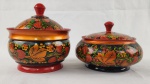 Conjunto composto por duas caixas com tampa, executadas em madeira torneada e ricamente decoradas com pintura manual em padrão floral policromado, com predominância de tons vermelho, dourado, verde e fundo escuro, característico do estilo russo Khokhloma. Peças de forte apelo decorativo, com acabamento envernizado e puxadores superiores. Medidas: aprox. 17 x 15 cm e 12 x 18 cm. 