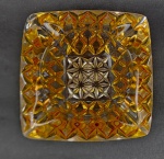 Cinzeiro em vidro prensado na tonalidade âmbar, de formato quadrado com bordas facetadas e rica decoração geométrica em relevo. Medidas: 6 x 13 cm.