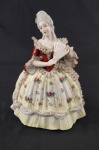 Escultura em porcelana representando dama de época, com traje estilo Rococó, ricamente detalhada, com vestido floral, rendados aplicados e leque nas mãos. Pintura policromada delicada. Medidas: 13 x 12 x 11 cm. Apresenta bicados.