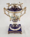 Ânfora ornamental em porcelana esmaltada policromada, decorada com reservas figurativas de inspiração rococó, representando personagens em cena campestre. Corpo ricamente adornado com relevos, esmaltação em azul-cobalto e detalhes dourados. Base quadrangular ornada e alças vazadas de desenho sinuoso. Apresenta restauro e ausência parcial de elemento decorativo em uma das alças. Altura: 24 cm.