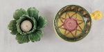 Duas peças; A)Delicado castiçal em cerâmica esmaltada modelada em forma de flor de repolho, com pétalas em relevo e esmaltação em tonalidades de verde. Peça de caráter decorativo, inspirada na tradicional cerâmica naturalista europeia. Medidas: 5 x 17 x 12 cm.B) Pequena tigela em cerâmica esmaltada, decorada manualmente com composição geométrica policromada em tons de amarelo, verde, marrom e rosa. Apresenta alça lateral integrada e decoração de inspiração artesanal, lembrando produções mediterrâneas e latino-americanas.Medidas: 6 x 13 cm.