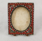 Miniatura - Delicada porta-retrato em metal de cloisonné vazado, decorada com esmaltação vermelha e ornatos geométricos de inspiração oriental. Medidas: 6 x 4,5 cm.