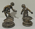 Par de tinteiros em metal espessurado a prata, em estilo Rococó, ornamentados por esculturas de querubins em relevo, representados em diferentes poses sobre bases ricamente decoradas com volutas, frisos e elementos florais, conjunto de mesa de excepcional apelo decorativo, inspirado na arte europeia do século XIX. Apresentam marcas do tempo e desgastes. Medindo: 20 x 23 cm e 17 x 20 cm.