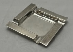 PRATA. Cinzeiro em prata argentina de teor 925 mls, de formato quadrangular, com quatro apoios para cigarro, centro liso e bordas largas com acabamento finamente guilhochado de inspiração Art Déco. Peso; 65 gramas.