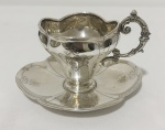PRATA. Antiga xícara com pires em prata de teor 800 mls, de elegante formato, ornamentado por finas gravações e delicado monograma na face frontal. Alça vazada ricamente trabalhada em volutas e pires de contorno recortado, decorado com filetes e motivos florais gravados. Peso total: 106 gramas.