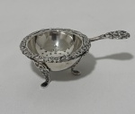 Antigo coador de chá em metal espessurado à prata inglesa, composto por coador e suporte de descanso. Coador de formato semiesférico, ricamente ornamentado em relevo com motivos vegetalistas, borda moldurada e cabo decorado em volutas. Apoiado sobre três pés estilizados, acompanhado de recipiente de apoio com borda ornamentada. Medidas: 10 cm, peso: 69 gramas
