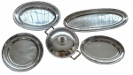 Wolff. Baixela em metal espessurado a prata, composta por cinco peças, sendo uma sopeira com tampa e alças laterais, três travessas ovais, uma travessa redonda. Bordas cinzeladas com delicado friso gravado. Medidas: sopeira 22 x 33,5 cm; travessas 33,5 cm, 37,5 x 26 cm, 42 x 31 cm e 60 x 23 cm.