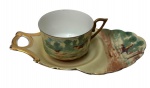 J. D. Moreira & Cia. Pernambuco. Antiga xícara de coleção em porcelana, acompanhada de Tea & Toast Tray (prato de apoio para chá e torradas), finamente decorada com cena campestre e pássaros em policromia, sobre fundo em tons suaves. Alça moldada, bordas recortadas e acabamento filetados a ouro. Carimbo do importador no verso. Medidas: 21 x 11,5 x 6 cm.