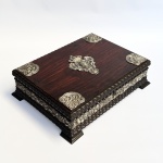 Caixa em jacarandá com aplicações em prata de lei ricamente cinzelada com motivos florais e brasão português ao centro. Interior forrado em veludo vermelho com dois estágios. Acompanha chave. Med 29 x 22 x 8 cm.