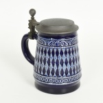 LOUÇA ALEMÃ, um (1) tankard na tonalidade azul cobalto com barrado relevado em treliças peroladas e volutas, tampa em pewter, marca parcialmente legível em cavo na base, medindo 14 cm altura, vide foto.