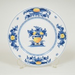 PORCELANA PORTUGUESA, um (1) prato decorativo, ornado com vaso de flores nas tonalidades azul e amarela sobre fundo branco, marca VISTA ALEGRE (fundada em 1824, pelo negociante, lavrador, industrial e político, José Ferreira Pinto Basto, através de alvará concedido por D. João VI, tendo seu fundador associado a fábrica os seus 15 filhos, ficando a sociedade denominada Ferreira Pinto & Filhos; cinco anos mais tarde recebe o título real pelo reconhecimento de sua arte e sucesso, ficando conhecida como Real Fábrica da Vista Alegre, começou no fabrico de vidros enquanto se empenhava em esforços para a produção de porcelana, os primeiros ensaios conseguidos foram imperfeitos denominado loiça de pó de pedra, através de boas relações com a fábrica francesa de Sevres conseguiu as receitas para o fabrico de porcelana, somente em 1832, por sorte, é encontrado um jazigo de caulim situado em Aveiro, próximo a Ílhavo, a partir desta data o fabrico de porcelana foi se aperfeiçoando em Portugal, 1851 expôs com sucesso no Palácio de Cristal em Londres; 1852 D. Fernando II visita a fábrica e encomenda uma baixela completa; 1867 recebeu sua primeira premiação na Exposição Universal de Paris; 1880 interrompe a produção de vidros para se dedicar exclusivamente a porcelana; 1964 inauguração do Museu Histórico da Vista Alegre; ainda em funcionamento), medindo 21 cm  diâmetro, vide foto.