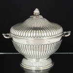 METAL PRATEADO, uma (1) fruteira de pé alto com tampa ou centro de mesa, ornamentação relevada em caneluras, pega encimada por morango, alça relevada com terminações em concha, sem marca de manufatura aparente, medindo 25 cm diâmetro mais as alças e 26 cm altura, sinais de uso, vide foto.