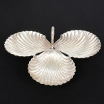 METAL PRATEADO, uma (1) petisqueira no formato de três conchas bivalves unidas formando um trevo, marca EBERLE (Abramo Eberle 1880, Monte Magrè 1945, Caxias do Sul, veio com uma das primeiras levas de imigrantes italianos para o Brasil em 1884 junto com seus pais Giuzeppe Eberle e Luigia Zanrosso, seu pai abriu uma pequena funilaria mas como tinha que trabalhar na lavoura quem cuidava era a mãe e o pequeno Abramo ajudava na funilaria; em 1896 quando o pai resolveu vender a funilaria para concentrar os esforços da família na lavoura Abramo, com 16 anos de idade, se propôs a comprá-la do pai, em 1918 já com o nome Abramo Eberle & Cia iniciou a fabricação de talheres e utensílios de mesa, morreu em 1945 com 65 anos de idade e os filhos deram continuidade aos negócios até 1984 quando venderam para o Grupo Zivi), alça relevada em guirlanda de tulipas com terminação em volutas, base trípode e esférica, medindo 19 x 16 cm, sinais de uso, vide foto.