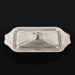AÇO INOX, uma (1) manteigueira com tampa, sem marca de manufatura aparente, medindo 19,5 x 10 cm, sinais de uso, vide foto.
