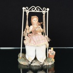 BRINQUEDO, uma (1) boneca sentada em cadeira de balanço, cabeça, membros superiores, corpo e pés,confeccionados em resina policromada, pernas em tecido com enchimento, cadeira de balanço em aramado vergado,soldado e com patina na tonalidade branca, assente sobre fundo em resina policromada, relevado em pedras e elementosvegetalistas, medindo 20,5 cm altura, pequenas falhas e perdas, possivelmente de origem italiana, vide foto.