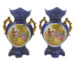 Par de solifleurs no estilo Sèvres, em azul cobalto com alças laterais com douração total; galeria central decorada com colorida cena galante; em porcelana pintada à mão (assinatura do artista no fundo). Medidas: 18 x 15 cm.