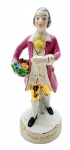 VIEIRA DE CASTRO RIO - Antigo bibelô com a representação de uma figura masculina em trajes de época segurando um buquê de flores; em porcelana multicolorida e com detalhes em dourado. Medidas: 20 x 8 cm. Objeto segurado pela mão esquerda com dano.