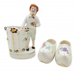 CSA PEDREIRA / SANTANA - 02 bibelôs em porcelana, sendo um representando uma figura masculina em trajes de época, da manufatura CSA Pedreira, e um par de tamancos no estilo holandês, da manufatura Santana; decorados com coloridos padrões florais e detalhes em dourado. Medidas: 11 x 7 cm; 8 x 4 cm. Um dos tamancos com ínfimo bicado na borda.