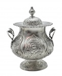 P COROADO - Potiche em prata de lei portuguesa contrastada, com linha em ziguezague (teste da burilada), peso: 454 g, séc. XIX; ricamente cinzelado em relevo com padrões florais, volutas e folhagens; alças laterais decoradas com padrões fitomórficos; base pedestal acompanha o padrão ornamental do restante da peça. Nota: A burilada (ziguezague) serve como uma assinatura histórica de época; antes dos métodos químicos modernos, o ensaiador da contrastaria usava uma ferramenta chamada buril para raspar uma pequena porção de metal da peça e testar o seu teor de pureza; somente depois disso ele aprovava e carimbava. Essa raspagem intencional deixava um sulco característico em forma de ziguezague. Medidas: 19 x 16 cm. Tampa com ínfimos amassados.