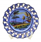 Medalhão em cerâmica esmaltada portuguesa, moldado com detalhes ondulados e orgânicos vazados na borda, decorado à mão com coloridos padrões fitomórficos e cenário com estrada e casebre em sua galeria central. Numerado. Medidas: 36 cm (diâm.). Com sinais de restauro na borda.