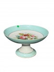 PORCELANA, uma (1) fruteira de pé confeccionado em porcelana na tonalidade branca, decorada por flores e folhagens em policromia e espessa faixa na tonalidade azul com filetes em ouro, apresentando leves falhas na douração e bicado na borda, medindo 12x21,5cm.