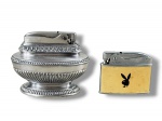 METAL PRATEADO, lote com dois (2) isqueiros, sendo: A) RONSON, 1 de mesa, confeccionado em metal espessurado a prata cinzelado em gomos, perolados e folhagens, necessita revisão, medindo 6,5x8x4,5cm e B) PLAYBOY, 1 confeccionado em metal prateado com decoração na tonalidade creme e símbolo da PLAYBOY, medindo 4,5x5,5cm, necessita revisão.
