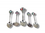 METAL PRATEADO, cinco (5) colheres souvenir de coleção, confeccionadas em metal espessurado, com cabos ornamentados e terminais decorados com brasões, inscrições e aplicações esmaltadas coloridas, representando localidades turísticas europeias, incluindo Interlaken, Hamburg, Suíça e outras.