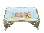 PORCELANA, um (1) saleiro confeccionado em porcelana na tonalidade branca, com pintura na cor azul e decoraçao de flores e folhagens em policromia assinado REDA, apoiado sobre estrutura em metal com patina envelhecida e sinais de oxidação, medindo 3,5x9x7cm.