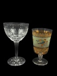 CRISTAL e VIDRO, lote com duas (2) taças, sendo: A) 1 para vinho confeccionada em cristal translucido com lapidação de caneluras e formas geométricas em baixo relevo, haste facetada, base circular com fundo estrelado, medindo 15,5x9cm e B) 1 para água confeccionada em vidro translúcido com vestígios de douração e pintura de pássaros e elementos florais, medindo 13,5x7,5cm, pequeno defeito de fabricação na base.